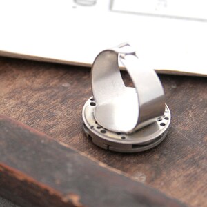 May include: A silver ring with a circular design featuring a gear-like pattern. The ring is made of metal and has a smooth, polished finish.