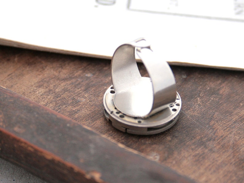 May include: A silver ring with a circular design featuring a gear-like pattern. The ring is made of metal and has a smooth, polished finish.