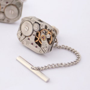 May include: Silver tie tack with a silver chain and a decorative watch movement design. The watch movement is visible through a clear glass cover.