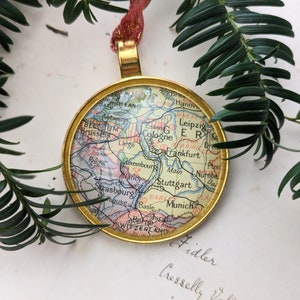 以下が含まれることがあります： ヨーロッパのヴィンテージ地図をあしらった、丸い金色のクリスマスオーナメント。ケルン、フランクフルト、シュトゥットガルトなどの都市が地図に表示されています。赤と金のリボンで吊り下げられ、背景には緑の葉があります。