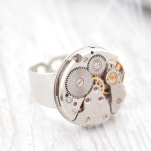 May include: A silver ring with a circular watch face featuring exposed gears and a silver band.