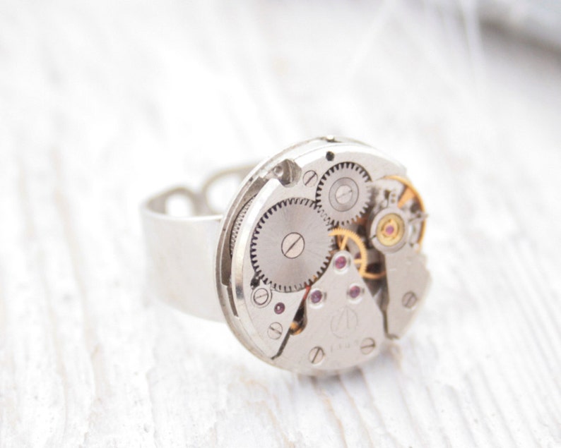 May include: A silver ring with a circular watch face featuring exposed gears and a silver band.