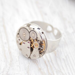 May include: A silver ring with a circular watch face featuring exposed gears and a gold-colored winding stem. The ring is made of metal and has a heart-shaped cutout on the band.