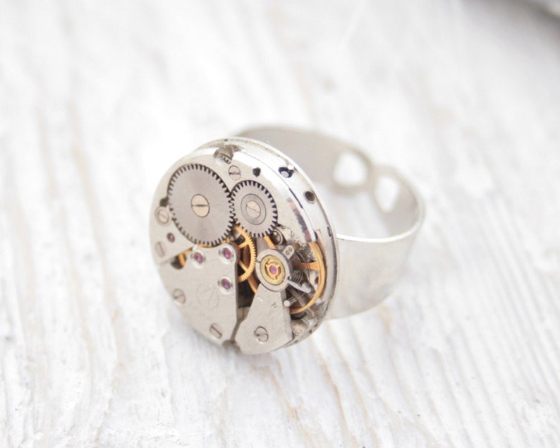 May include: A silver ring with a circular watch face featuring exposed gears and a gold-colored winding stem. The ring is made of metal and has a heart-shaped cutout on the band.