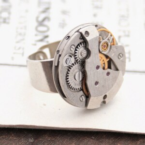 May include: A silver ring with a circular watch face featuring exposed gears and a small ruby jewel. The watch face is attached to a silver band.