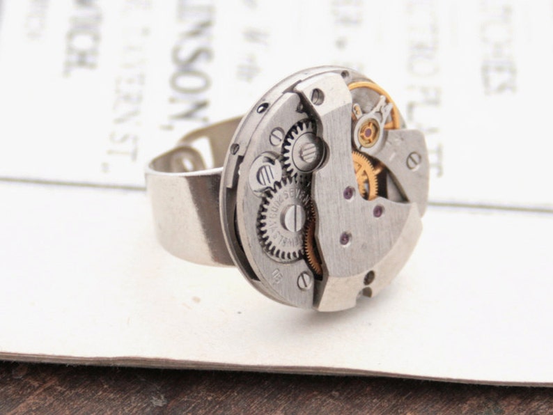 May include: A silver ring with a circular watch face featuring exposed gears and a small ruby jewel. The watch face is attached to a silver band.