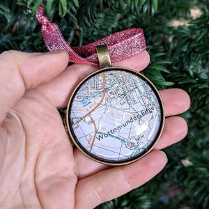 May include: A round Christmas ornament with a map of Wotton-under-Edge, England, encased in a bronze-colored metal frame. The ornament is suspended by a red ribbon. The map includes street names, roads, and the B4050 and B4060 roads.