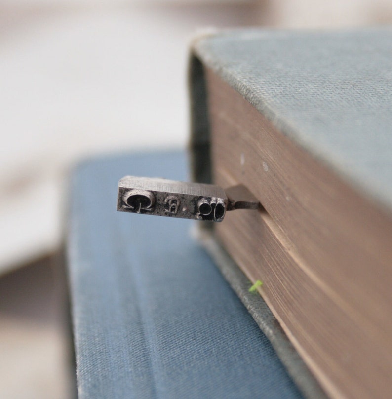 May include: A close-up of a silver bookmark with the letters "Q", "S", and "2" on it. The bookmark is sticking out of the edge of a blue book.