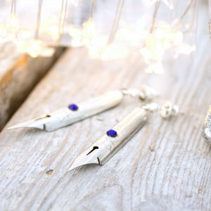 May include: A pair of silver earrings shaped like calligraphy nibs with blue gemstone accents.