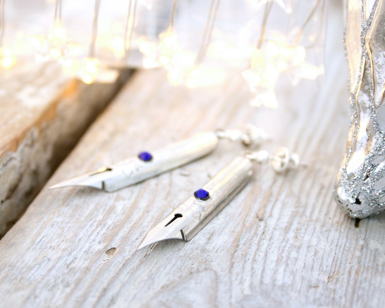 May include: A pair of silver earrings shaped like calligraphy nibs with blue gemstone accents.