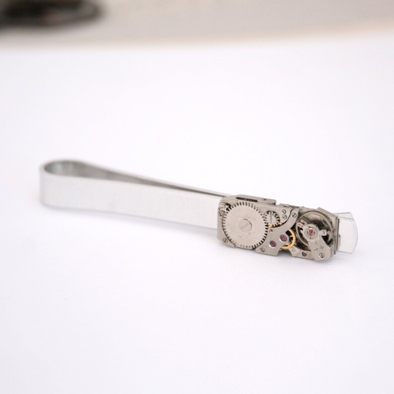 Puede incluir: Pinza de corbata plateada con un mecanismo de reloj visible. La pinza est&aacute; hecha de metal y tiene un acabado plateado. El mecanismo del reloj es visible a trav&eacute;s de una ventana transparente en la pinza.