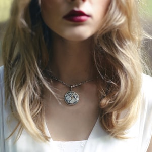 May include: A woman with long blonde hair wearing a white sleeveless top with a silver necklace featuring a round pendant with a clockwork design.