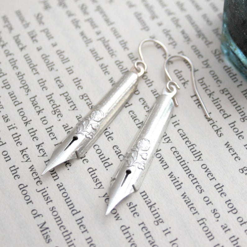 Puede incluir: Pendientes de plata con forma de plum&iacute;n vintage con dise&ntilde;os florales. Los pendientes se muestran en una p&aacute;gina de un libro.