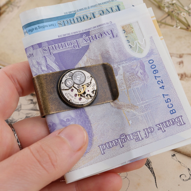 May include: A vintage-style money clip made of bronze-toned metal, holding several British banknotes. The clip features a circular watch mechanism with visible gears and jewels. The banknotes are visible, with the text "Twenty Pounds" and "Five Pounds".