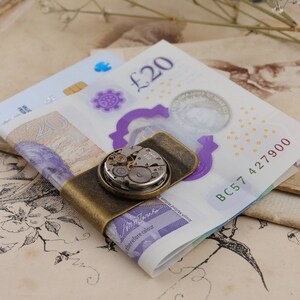 May include: A vintage-style money clip made of brass, holding a twenty-pound note and a credit card. The clip features a watch mechanism as a decorative element, showcasing intricate gears and metal components.
