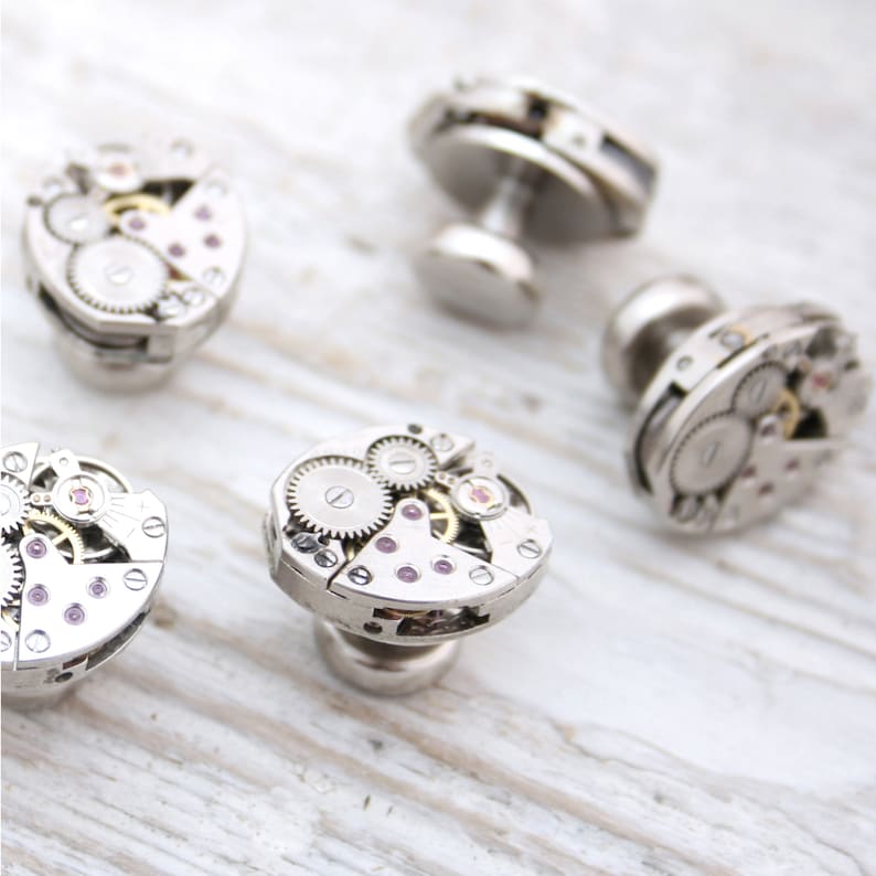 May include: Four silver cufflinks with intricate clockwork designs. Each cufflink features a different arrangement of gears and cogs, with some having small pink accents.
