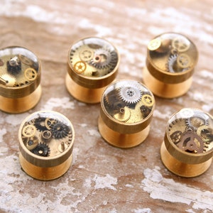 May include: Six gold-toned metal cufflinks with clear resin tops. Each cufflink features a different arrangement of small, gold-colored gears.