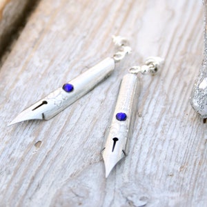 May include: A pair of silver earrings shaped like vintage dip pen nibs with blue gemstone accents.