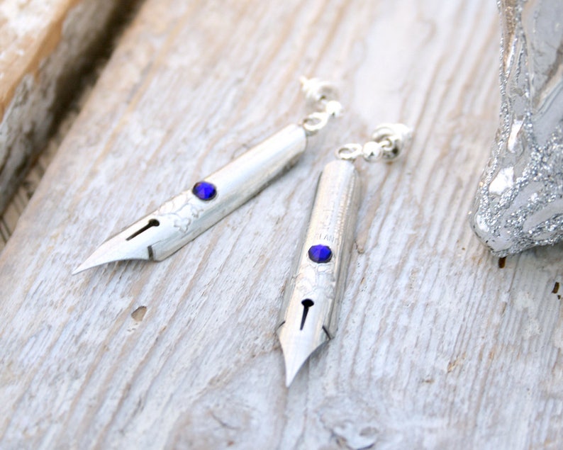May include: A pair of silver earrings shaped like vintage dip pen nibs with blue gemstone accents.