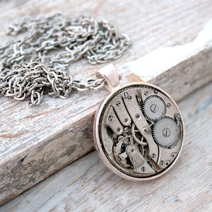 Può includere: Collana con pendente argentato con un pendente circolare con ingranaggi e meccanismi di orologio intricati. Il pendente è sospeso a una catena d'argento. La collana è esposta su una superficie di legno chiaro e invecchiato.