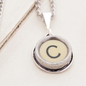 May include: A silver chain necklace with a pendant featuring a vintage typewriter key with the letter 'C' in black on a cream-colored background.