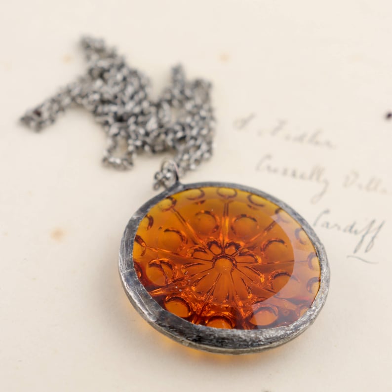 Puede incluir: Un collar de cadena de plata con un colgante redondo de vidrio &aacute;mbar. El colgante tiene un dise&ntilde;o detallado e intrincado de una flor.