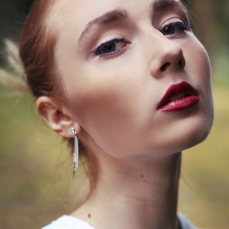 Puede incluir: Primer plano de la cara de una mujer con l&aacute;piz labial rojo y aretes largos de plata. Su cabello est&aacute; recogido hacia atr&aacute;s y est&aacute; mirando hacia un lado.