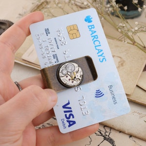 May include: A bronze-colored money clip holding a Barclays credit card and a Visa debit card. The clip features a circular watch mechanism design. The cards are held by a hand.