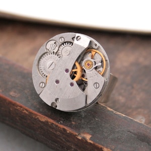 May include: A silver ring with a circular watch face featuring exposed gears and a gold-coloured winding mechanism.