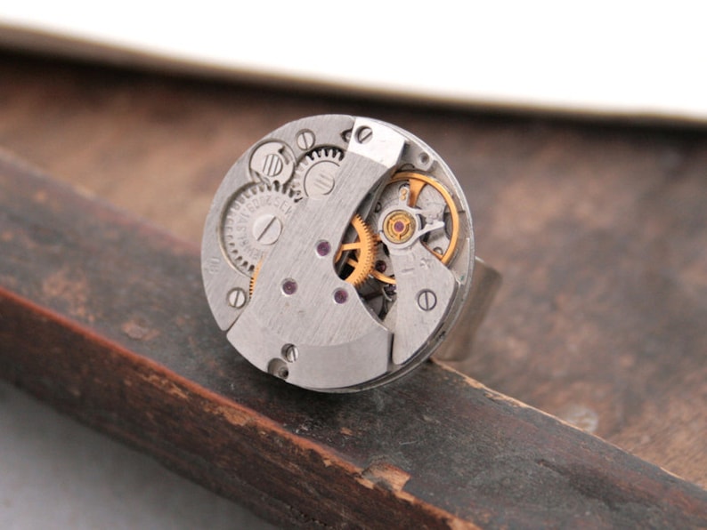 May include: A silver ring with a circular watch face featuring exposed gears and a gold-colored winding mechanism.