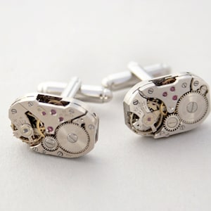 May include: A pair of silver cufflinks with a unique design featuring exposed watch gears and pink gemstones. The cufflinks are made of metal and have a polished finish.