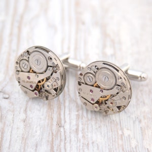 Könnte beinhalten: Ein Paar silberne Manschettenknöpfe mit dem komplizierten Innenleben einer Uhr. Die runden, metallischen Oberflächen zeigen Zahnräder, winzige Schrauben und rote Juwelen. Ein Manschettenknopf trägt die Aufschrift "15 Jewels".