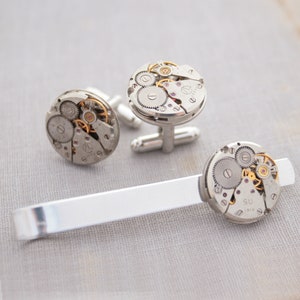 May include: A set of silver cufflinks and a tie bar featuring exposed watch gears. The cufflinks have a silver finish and the tie bar is silver with a polished finish.