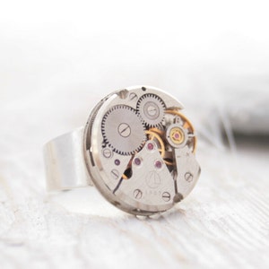 May include: A silver ring with a circular watch face featuring exposed gears and a small red jewel. The watch face is set into a silver band.