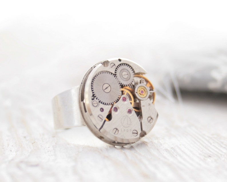 May include: A silver ring with a circular watch face featuring exposed gears and a small red jewel. The watch face is set into a silver band.