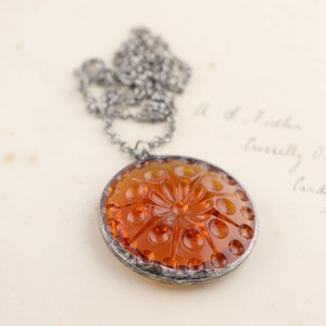 Puede incluir: Un collar de cadena de plata con un colgante de vidrio &aacute;mbar. El colgante es redondo y presenta un dise&ntilde;o floral en relieve.