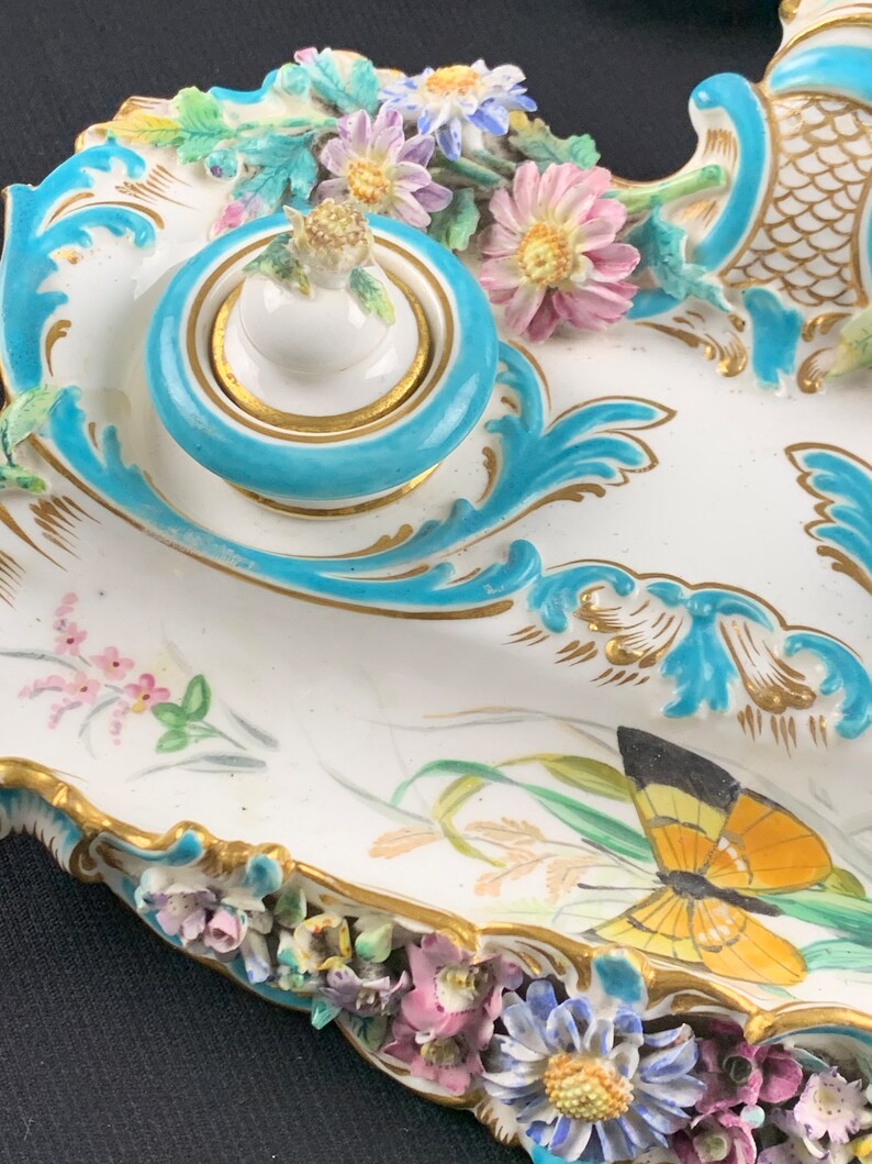 May include: A porcelain inkwell set with a blue and gold design. The inkwell is decorated with flowers and a butterfly.