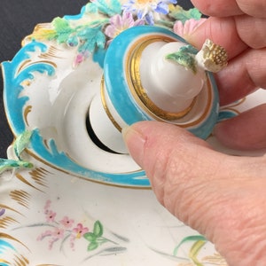 May include: A white porcelain inkwell with a blue and gold rim and a lid with a floral design. The inkwell is decorated with flowers and leaves in shades of pink, green, and yellow.