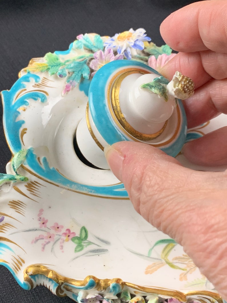 May include: A white porcelain inkwell with a blue and gold rim and a lid with a floral design. The inkwell is decorated with flowers and leaves in shades of pink, green, and yellow.
