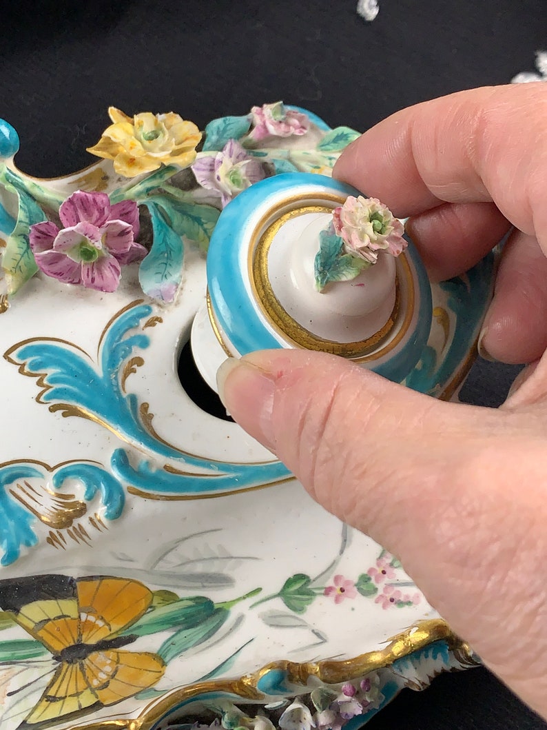 May include: A porcelain inkwell with a floral design and a butterfly. The inkwell has a blue and gold trim and a removable lid with a floral design.