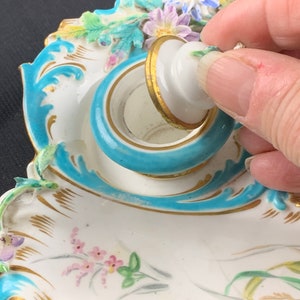 May include: A porcelain inkwell with a blue and gold rim and a lid with a floral design. The inkwell is decorated with painted flowers and leaves.