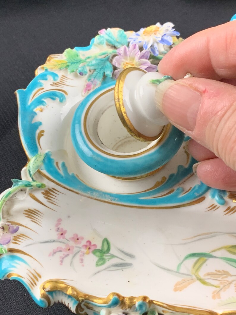 May include: A porcelain inkwell with a blue and gold rim and a lid with a floral design. The inkwell is decorated with painted flowers and leaves.