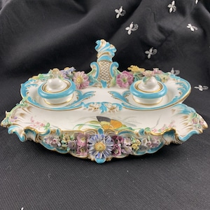 May include: A white porcelain inkwell with two inkwells and a central handle. The inkwells are decorated with blue and gold accents and the handle is decorated with a floral design. The inkwell is surrounded by a border of flowers and leaves.