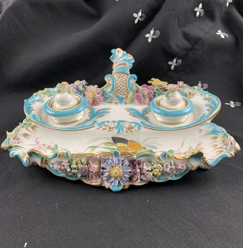 May include: A white porcelain inkwell with two inkwells and a central handle. The inkwells are decorated with blue and gold accents and the handle is decorated with a floral design. The inkwell is surrounded by a border of flowers and leaves.