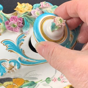 May include: A white porcelain inkwell with a blue and gold trim. The inkwell is decorated with flowers, butterflies, and leaves. The lid is blue with a gold trim and a white porcelain knob with a pink flower.