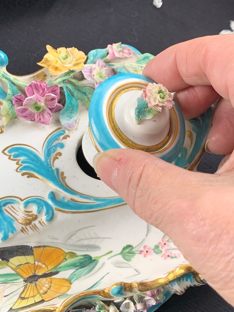May include: A white porcelain inkwell with a blue and gold trim. The inkwell is decorated with flowers, butterflies, and leaves. The lid is blue with a gold trim and a white porcelain knob with a pink flower.