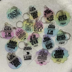 May include: A collection of colorful acrylic keychains with various book-themed quotes and designs. The keychains feature phrases like "Book Worm," "Dark Romance Reader," and "Books Over Bullsh*ts." Each keychain has a silver keyring.