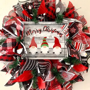 May include: A Christmas wreath with red, black, and white plaid ribbon, red and white buffalo plaid ribbon, and green pine sprigs. The wreath has a sign in the center that says "Merry Christmas" with three gnomes on it.