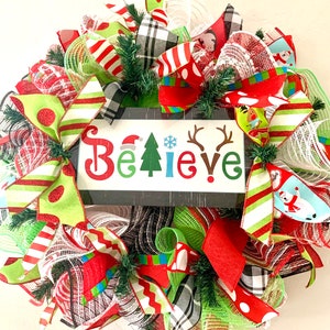 May include: A Christmas wreath with red, green, white, and black plaid ribbon, mesh, and polka dot fabric. The wreath has a sign that says "Believe" in colorful letters.