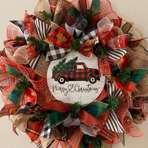 May include: A Christmas wreath with red, black, and white buffalo plaid ribbon, green and white plaid ribbon, and burlap ribbon. The wreath has a red truck with a Christmas tree in the back and the words "Merry Christmas" on a white sign.